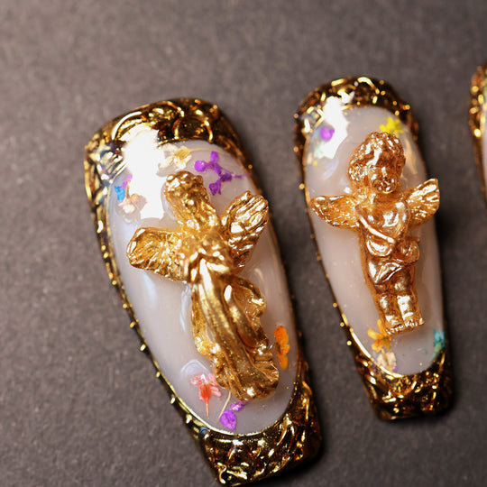 Angel Glow press-on nails in close-up, milky base with dried botanicals and ornate gold cherub charms