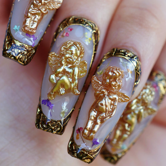 Angel Glow press-on nails with milky base, gold cherub charms and ornate borders, shown on hand