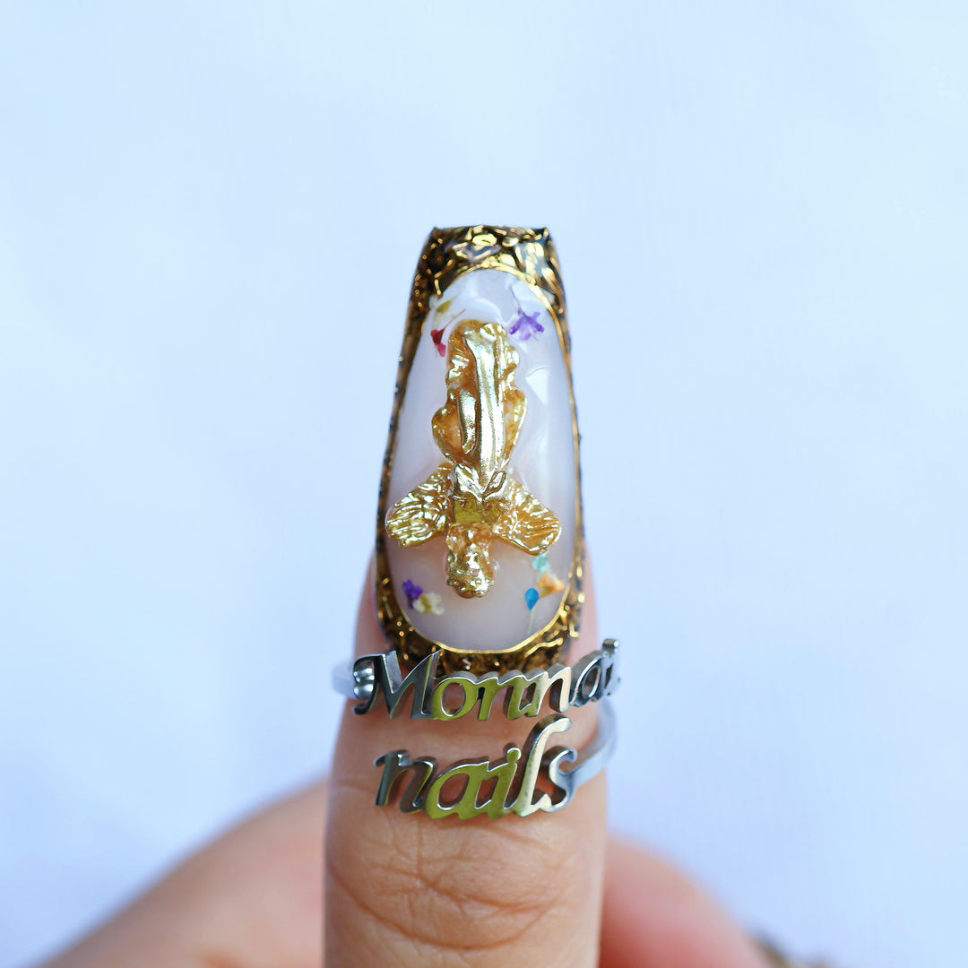 Angel Glow press-on nails worn on a finger, with milky base, gold cherub charm, floral details, and ornate gold border