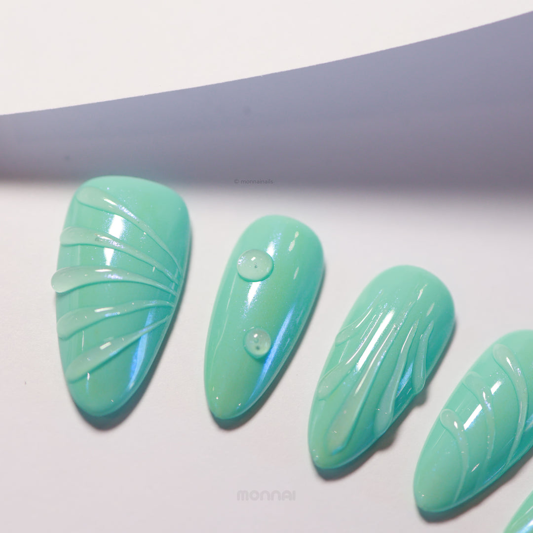 Aqua Glow press-on nails laid out with glossy teal bases, fine sculpted lines and droplet details