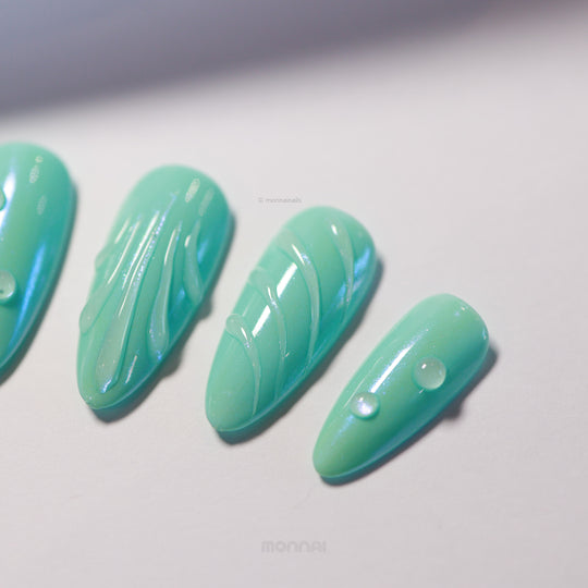 Aqua Glow press-on nails laid out in close-up, glossy teal with sculpted lines and droplet details