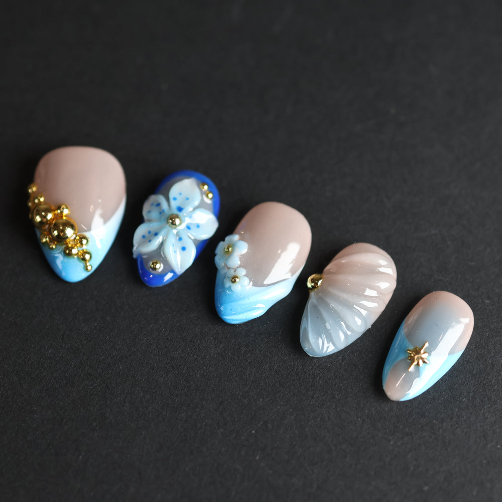 Aquamarine press-on nails laid out with nude bases, blue gradient tips, gold beads, and 3D floral details