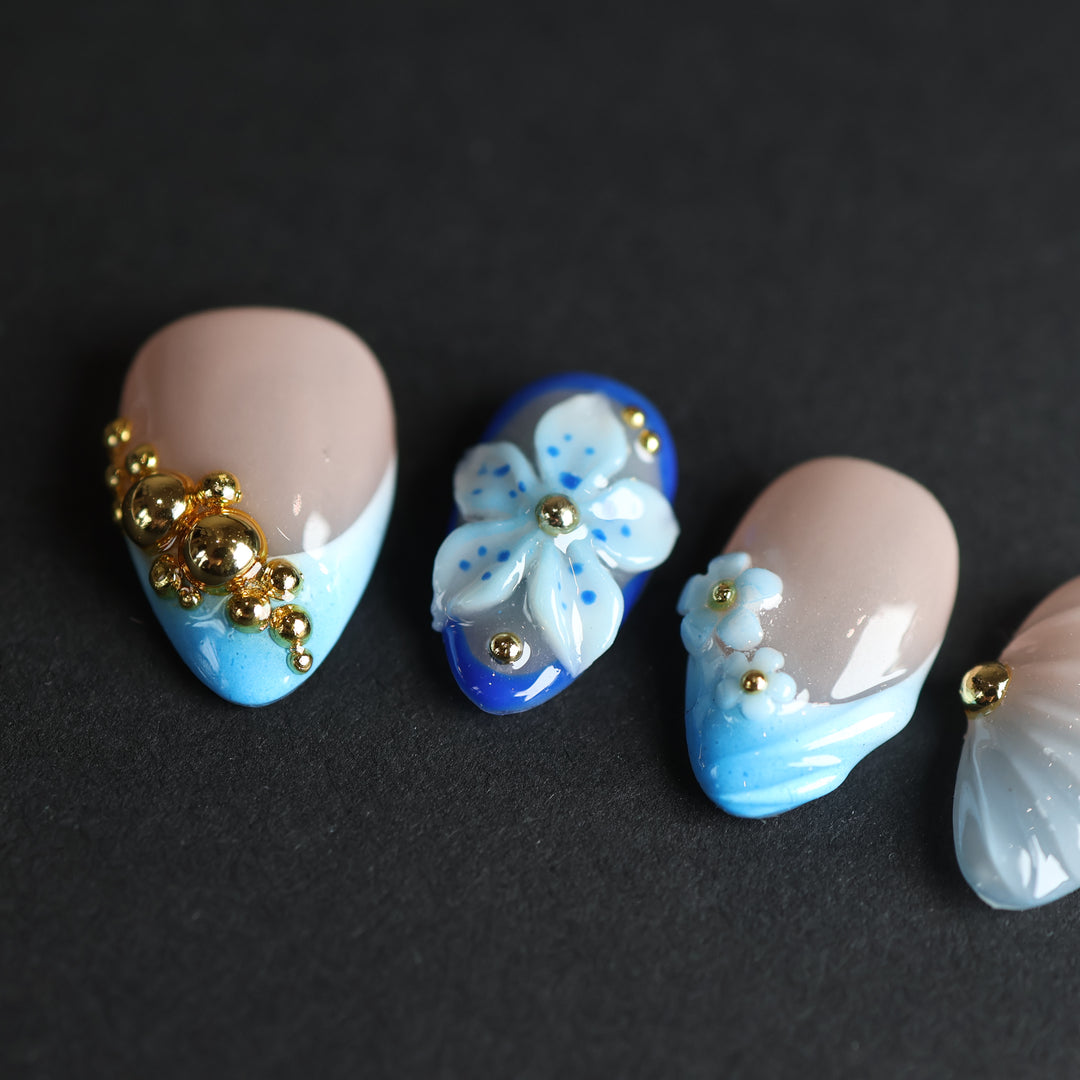 Aquamarine press-on nails with nude bases, blue gradient tips, 3D flowers, and gold beads laid out on black