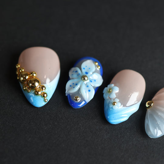 Aquamarine press-on nails with nude bases, blue gradient tips, 3D flowers, and gold beads laid out on black