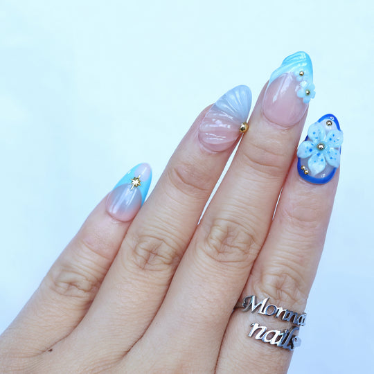 Aquamarine press-on nails on hand with blue gradient tips, 3D flowers, gold beads, and a seashell ridge design.