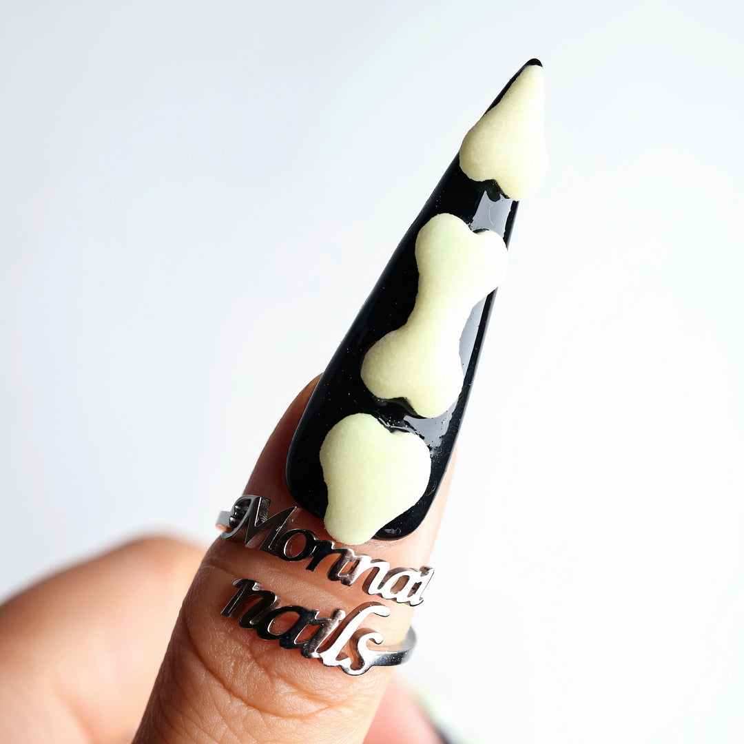 Bad to the Bone press-on nails shown on a hand, with black glossy tips and raised glow-in-the-dark bone details.