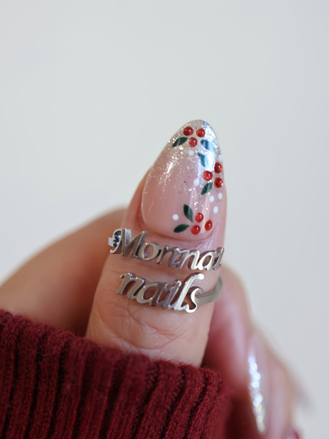 Berry Frost Christmas-themed press-on nails close-up worn on thumb with silver glitter, holly berries, leaves, and ruby rhinestones.