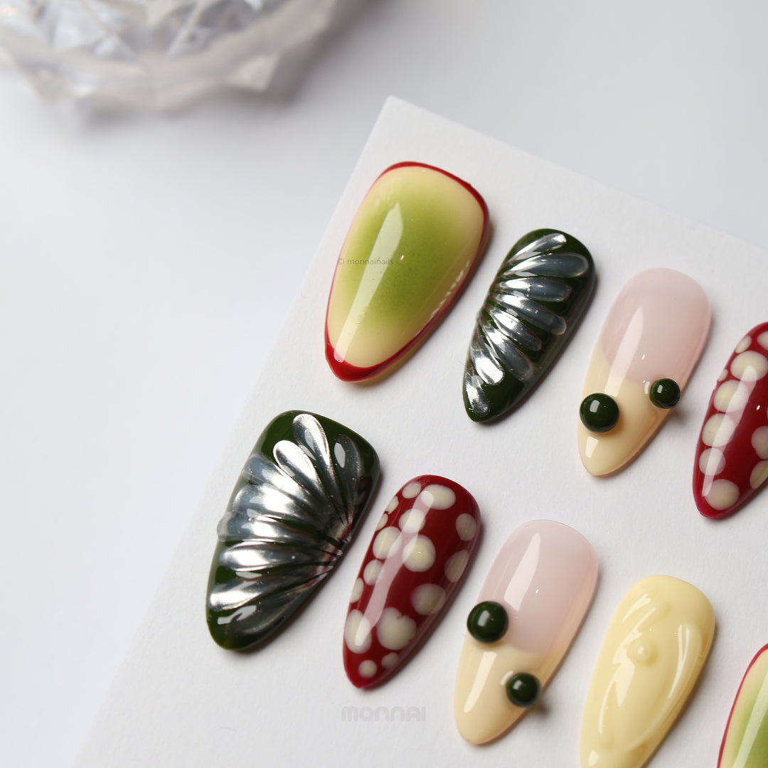 Berry press-on nails laid out, showing bold mixed designs with cream, nude, green, red, and glossy textured details.