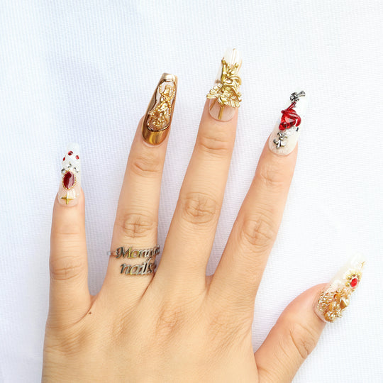 Blood Oath gothic press-on nails worn on hand with gold cherub charms, ruby-red jewels, and metallic Baroque detailing