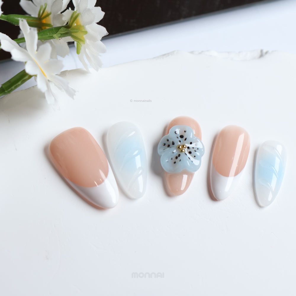 Blue Orchid press-on nails laid out, with soft nude, baby blue and French tip designs plus one 3D flower accent