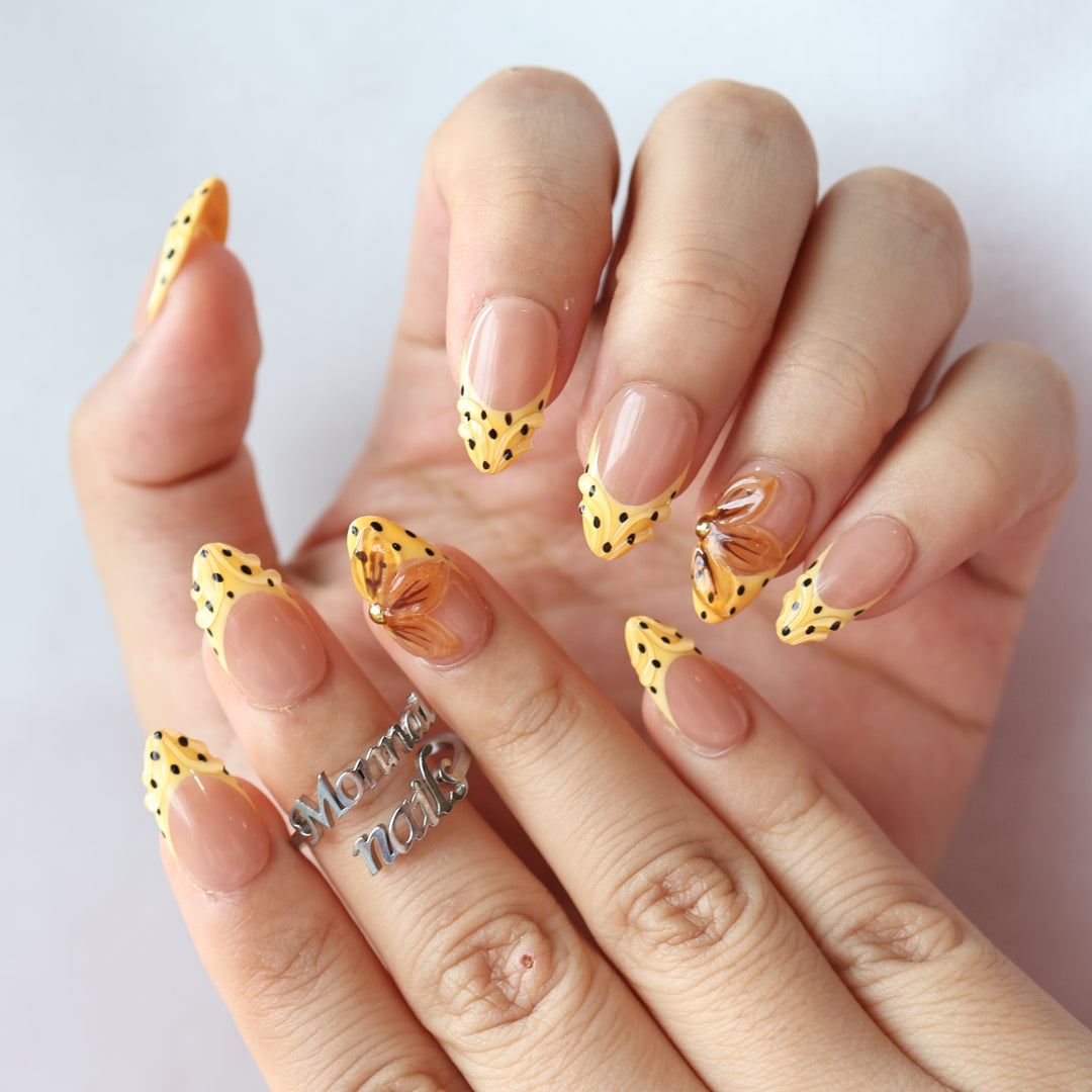 Caramel Dots press-on nails worn on hands, with butter yellow wavy French tips, black polka dots, and amber flower accents.