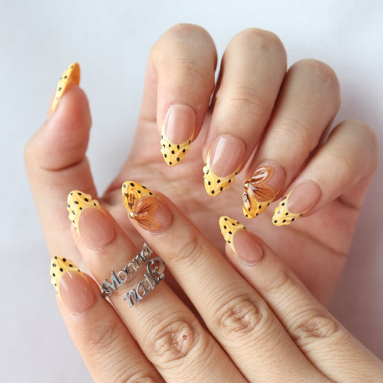 Caramel Dots press-on nails worn on hands, with butter yellow wavy French tips, black polka dots, and amber flower accents.
