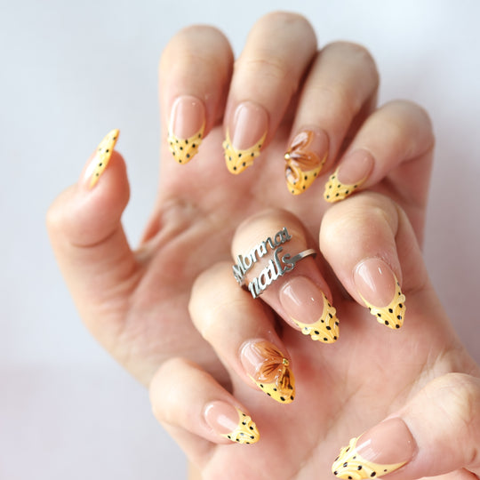 Caramel Dots press-on nails worn on hand with butter yellow wavy French tips, black polka dots, and translucent amber flower accents.