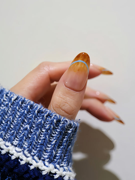 Caramel Mist press-on nails worn on hand, tortoiseshell tips and a crisp light blue arch with glossy nude base.