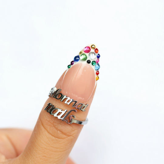 Carnival Lights press-on nails worn on a thumb, nude base with rainbow crystals and gold star accents