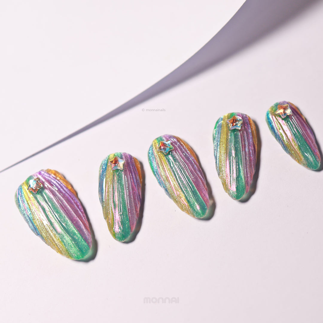 Cosmic Glow press-on nails laid out with 3D seashell texture, iridescent teal, pink and gold shimmer, each with a star rhinestone.