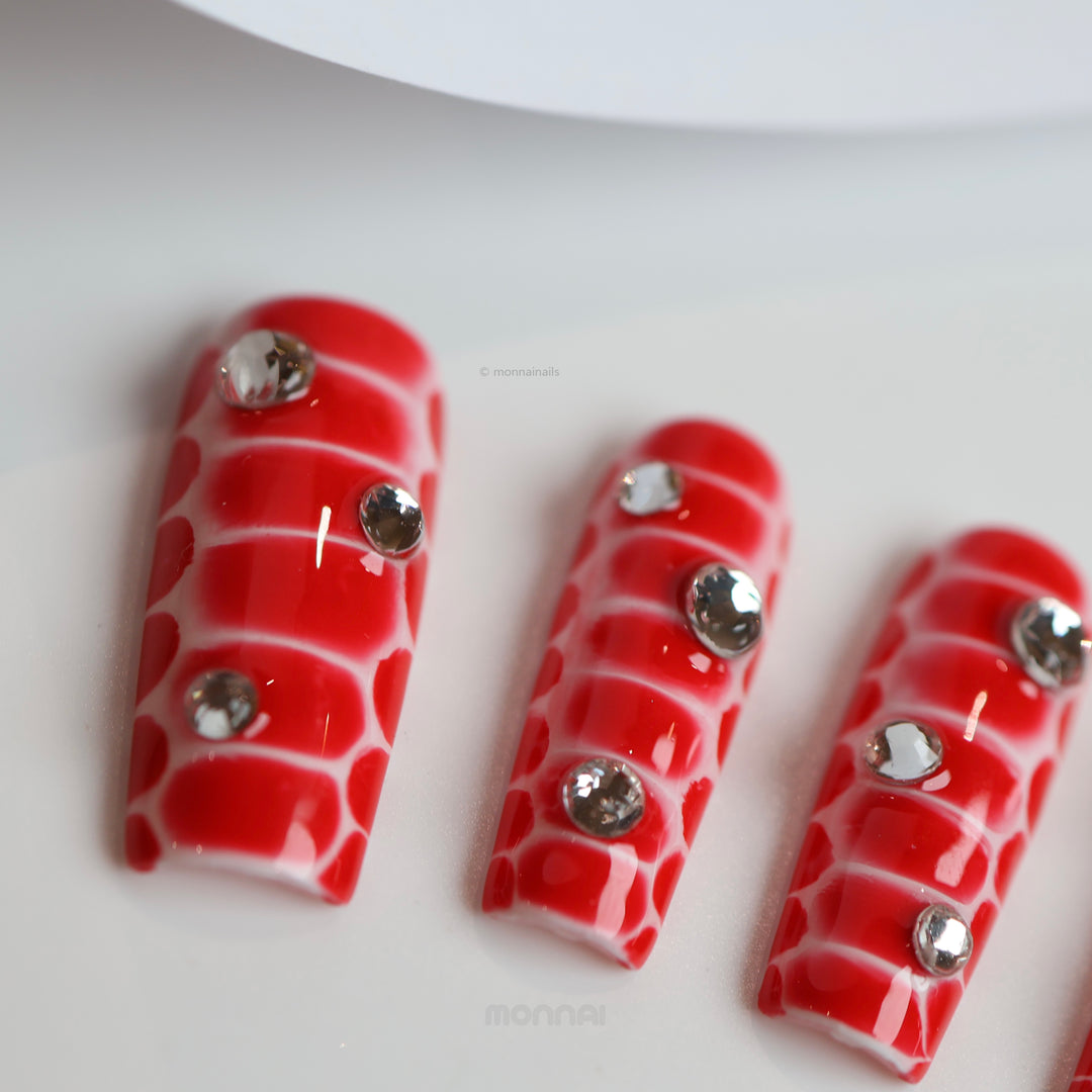 Crocodile Chic red press-on nails with sculpted crocodile texture and clear rhinestones, shown laid out in close-up.
