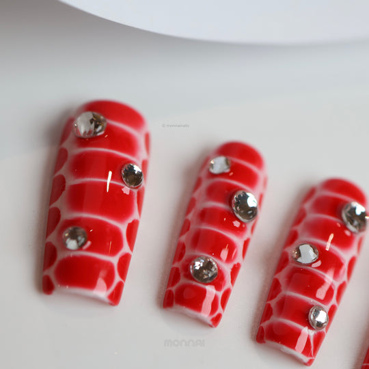 Crocodile Chic red press-on nails with sculpted crocodile texture and clear rhinestones, shown laid out in close-up.