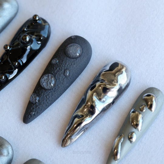 Dark Moon press-on nails laid out in close-up, with crater-like texture, raised gel droplets, and silver black metallic contrast.