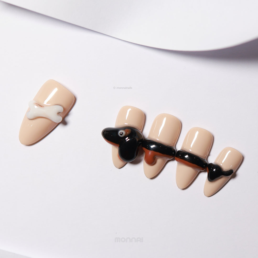 Doggy Delight press-on nails laid out in close-up, featuring a creamy nude base with a 3D dachshund motif and bone detail.