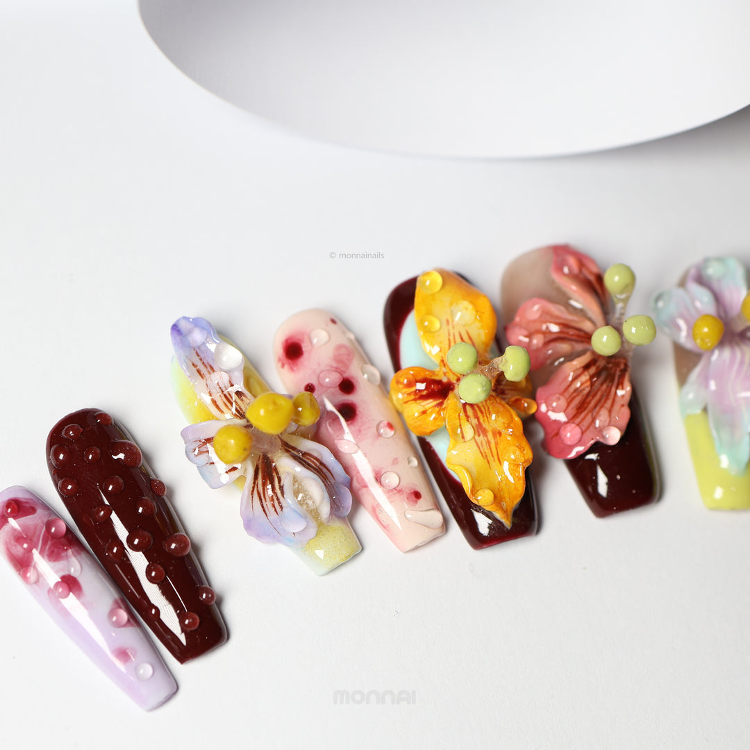 Dreamscape Blossoms press-on nails laid out in close-up, featuring botanical art, 3D petals, dewdrops, and rich ochre, bordeaux, and lavender tones.