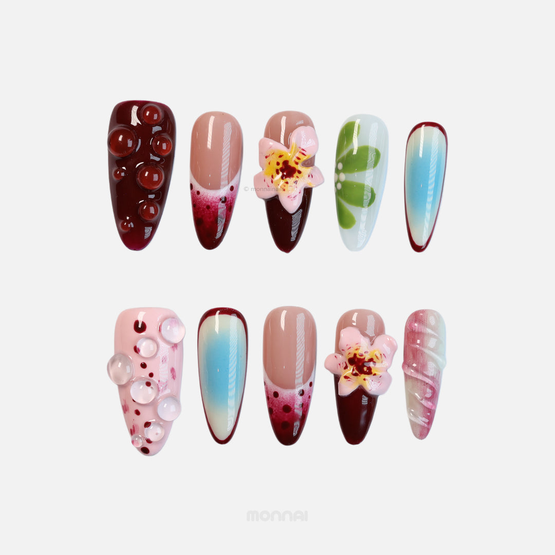 Floral Bliss press-on nails with cherry red, pastel, sky-blue aura gradients, floral 3D accents, and clear dewdrops laid out.