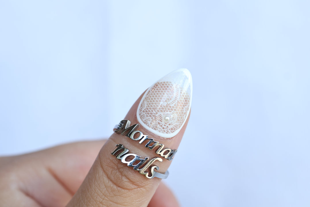Floral Veil press-on nails shown on a thumb, sheer white floral lace with a micro pearl accent and glossy finish.