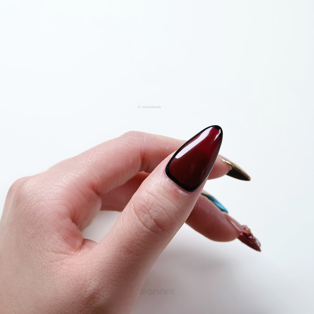 Gilded Rouge press-on nails worn on hand, deep wine red with glossy black outline and polished seasonal style.