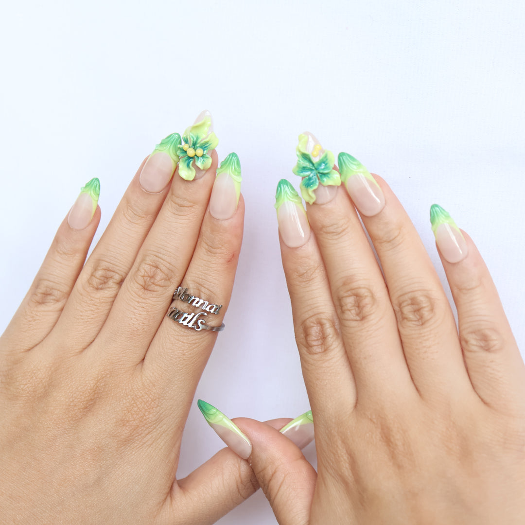 Green Bloom press-on nails worn on hands, sheer nude with green French tips and 3D floral accents.