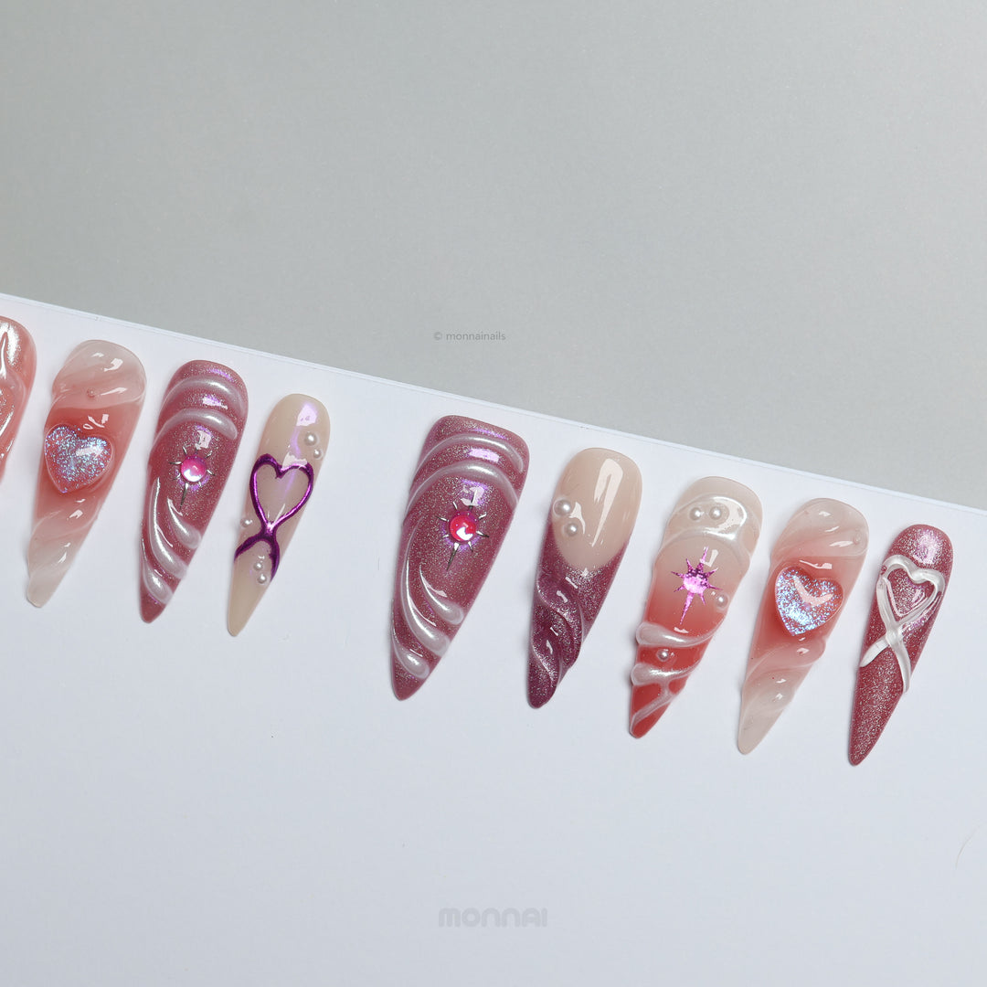 Heart’s Luster press-on nails laid out with pink glitter, glossy swirl details, and heart motifs on a soft nude base