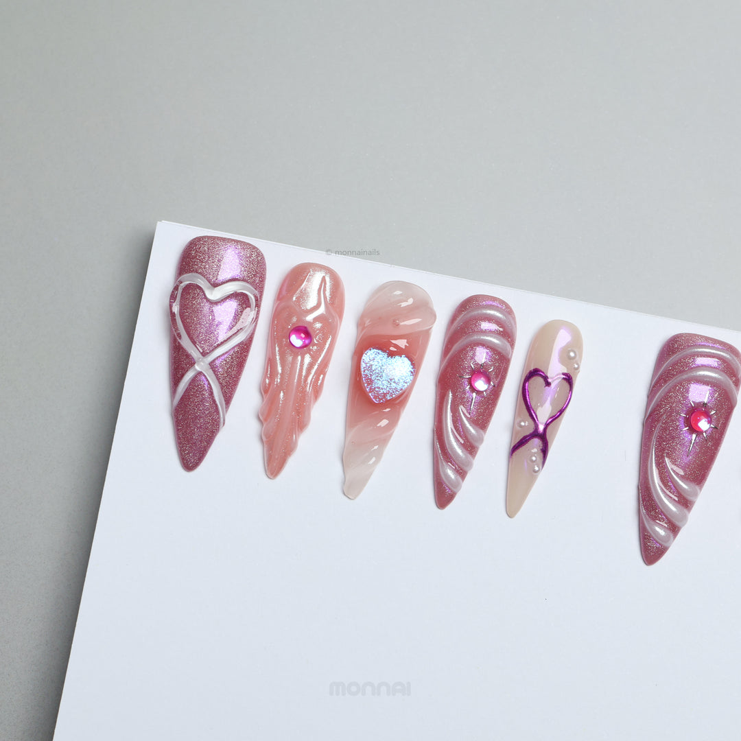 Heart’s Luster press-on nails laid out, with pink glitter, glossy heart motifs, sculpted swirls, and nude accents.