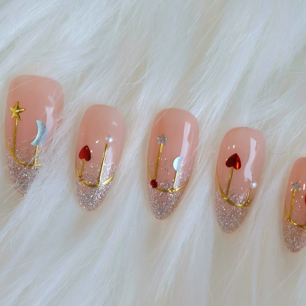 Holiday Glow press-on nails in close-up, soft pink with silver glitter French tips and gold star, moon, and heart accents.