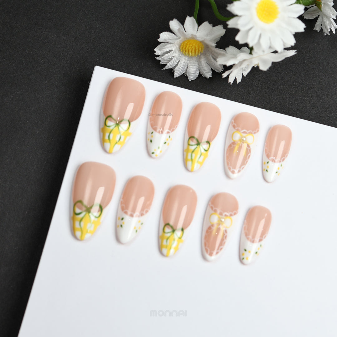 Lemon Lace press-on nails laid out with nude bases, white French tips, yellow gingham bows, and lace edging in a cottagecore style