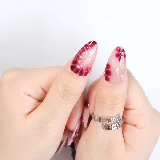 Lover’s Kiss press-on nails worn on hands, with sheer nude pink and deep burgundy floral details, silver beads, and glossy finish.