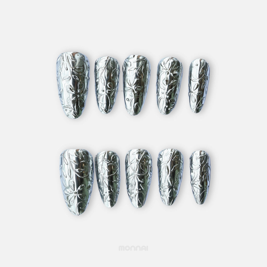 Metallic Bloom press-on nails with silver chrome floral engraving, shown laid out in a reflective close-up