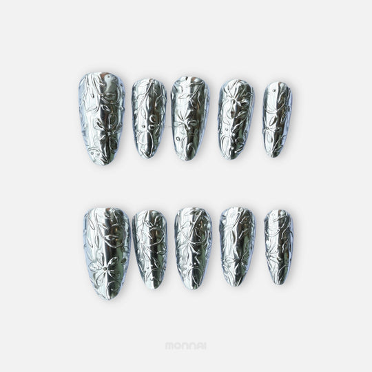 Metallic Bloom press-on nails with silver chrome floral engraving, shown laid out in a reflective close-up