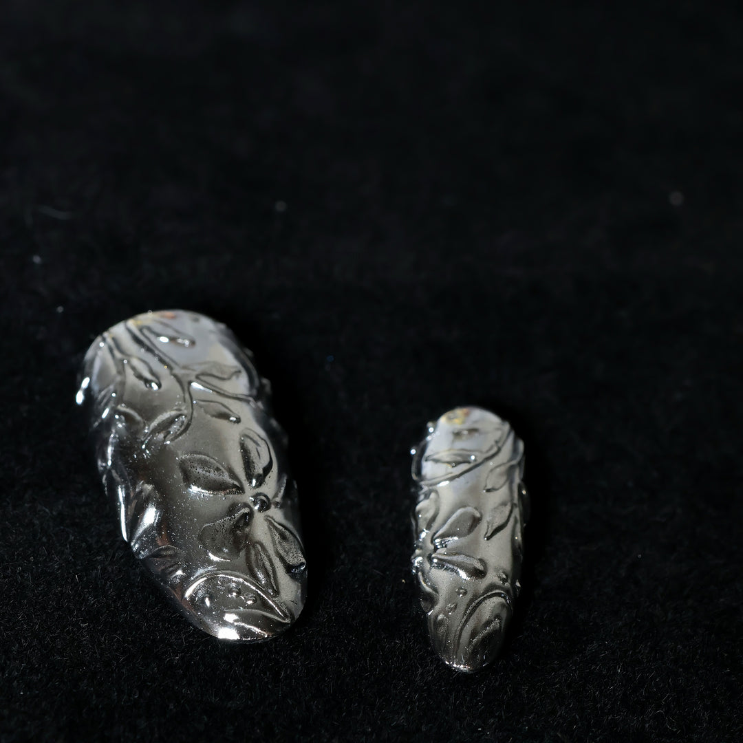 Metallic Bloom press-on nails in close-up, silver chrome with intricate floral engraving and raised sculptural linework.