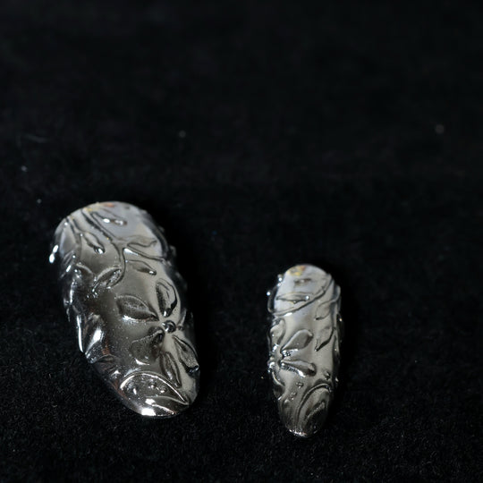Metallic Bloom press-on nails in close-up, silver chrome with intricate floral engraving and raised sculptural linework.