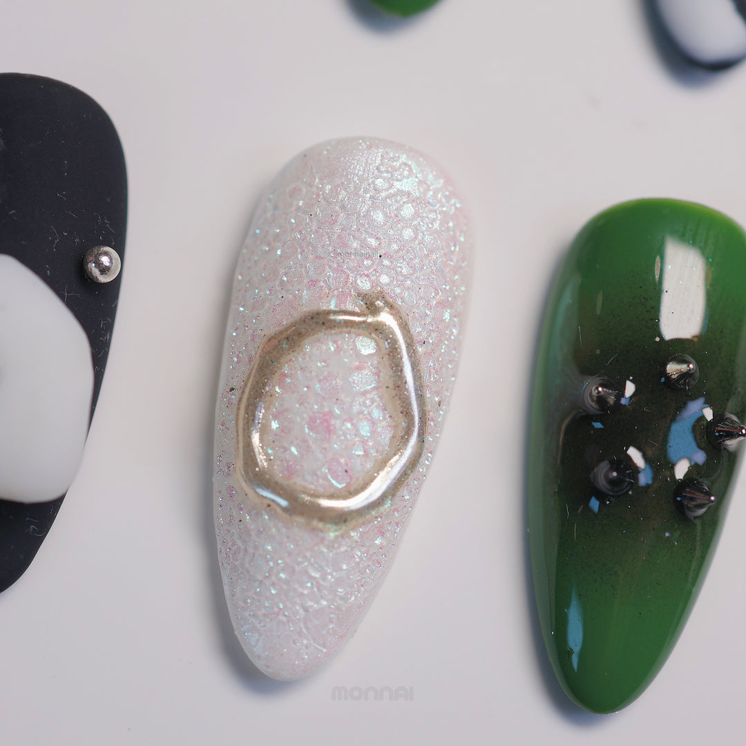 Metallic Mist press-on nails laid out in close-up, showing a sculptural iridescent nail with metallic accents and olive green tones.