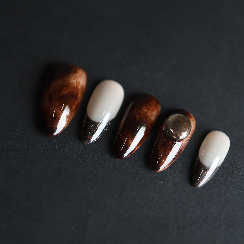 Molten Caramel press-on nails laid out with espresso cat-eye shimmer, nude bases, dark French tips, and a silver dome stud.