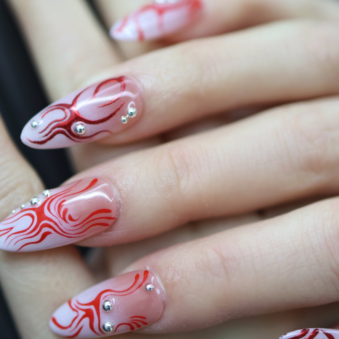 Neural Flux press-on nails on hand with red swirls, silver beads, and glossy sheer nude finish