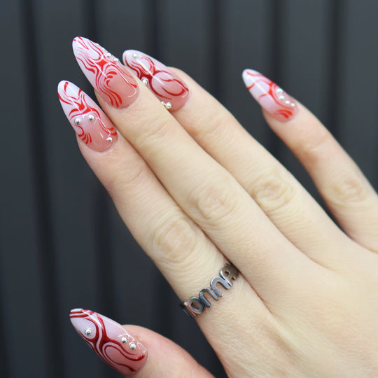 Neural Flux press-on nails on hand with red swirl art, sheer nude base, and silver beads