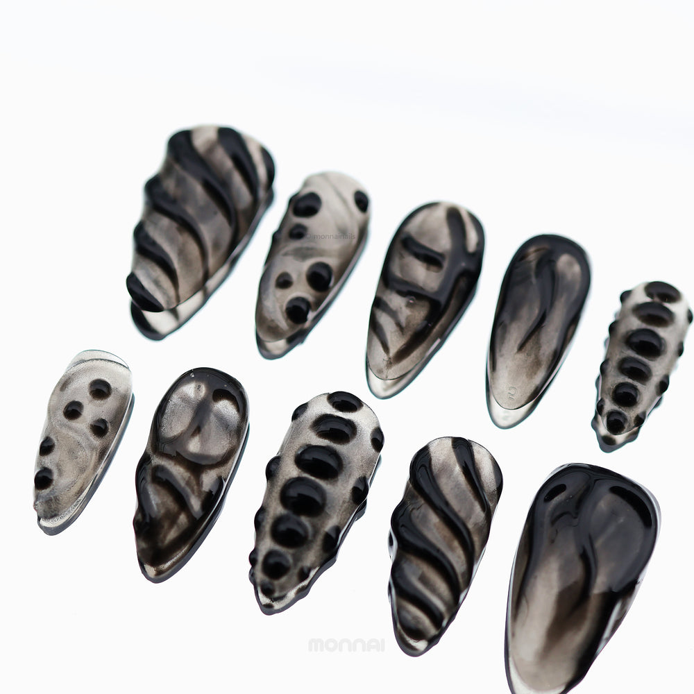 Obsidian Mist press-on nails laid out in close-up, with sheer nude bases, abstract black detailing, and glossy bead accents.