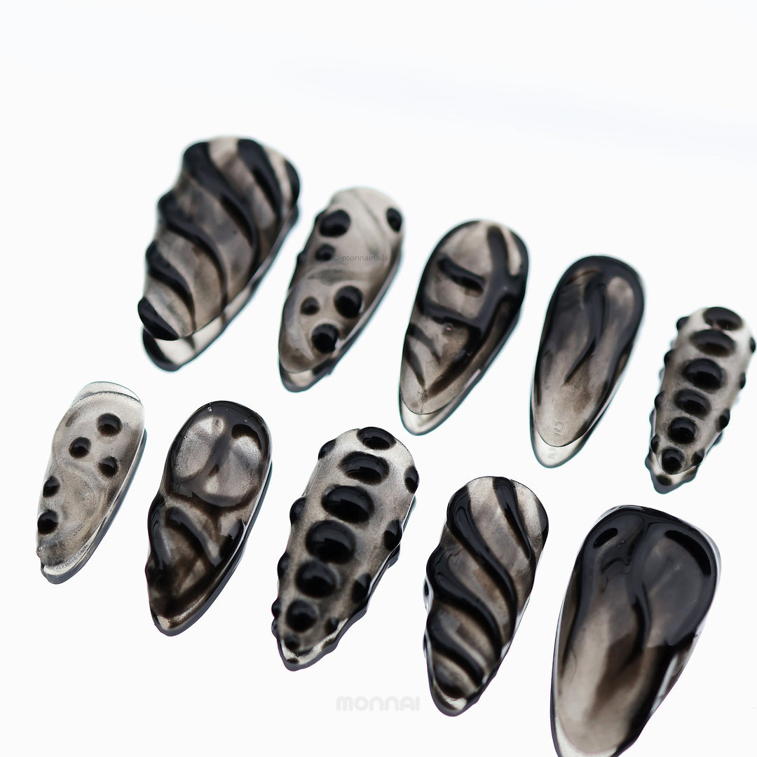 Obsidian Mist press-on nails laid out in close-up, with sheer nude bases, abstract black detailing, and glossy bead accents.