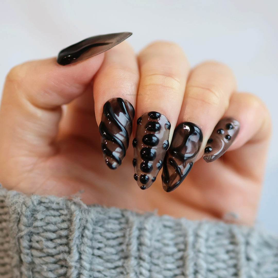 Obsidian Mist press-on nails on hand, sheer nude with black abstract gel lines and glossy bead accents