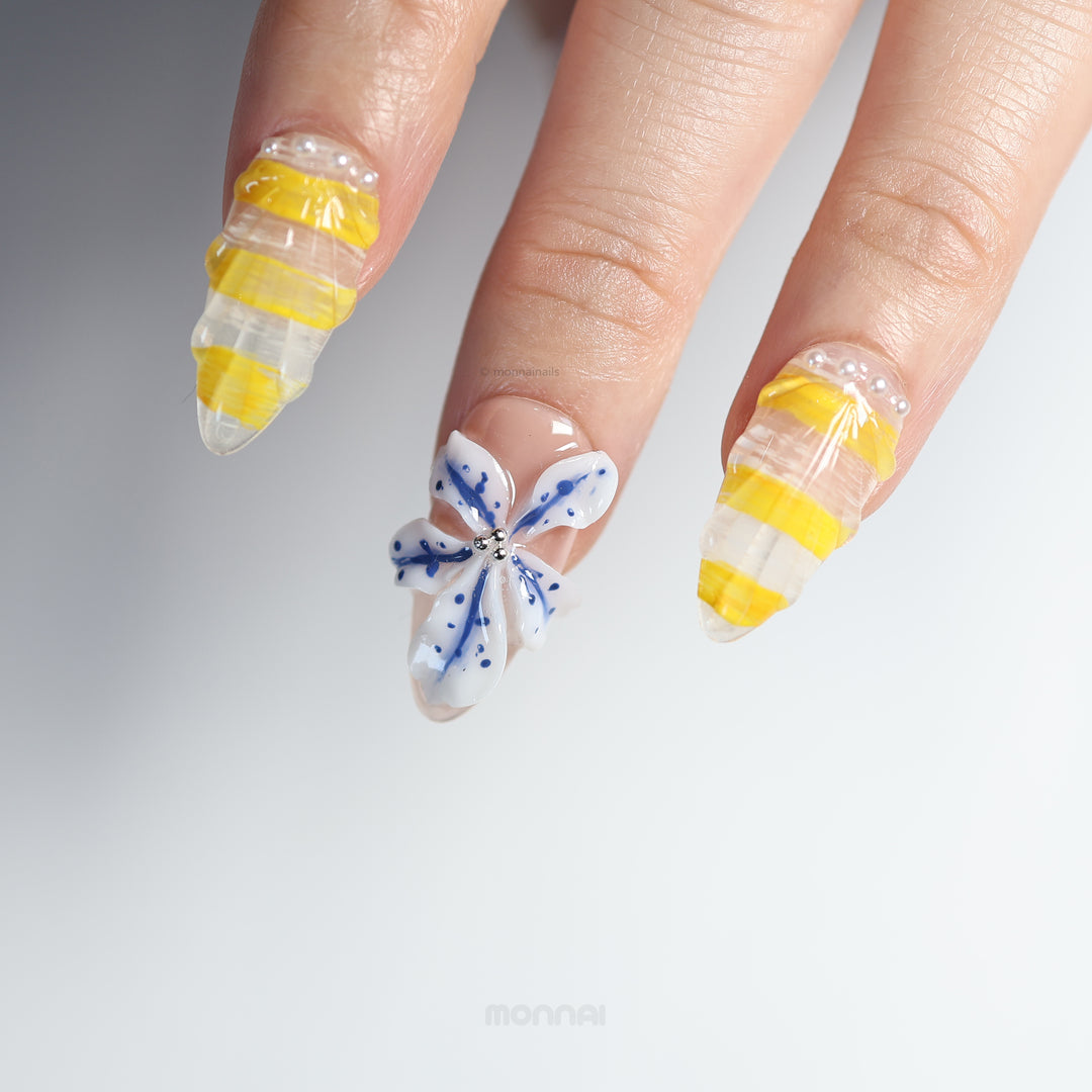 Ocean Bloom press-on nails on hand with glossy yellow stripes, sheer base, blue floral accent, and tiny bead details
