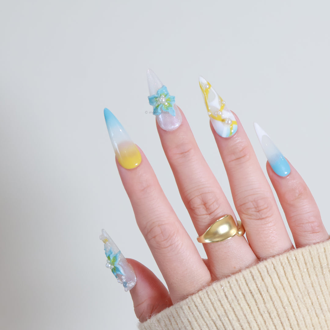 Ocean Breeze press-on nails worn on hand, with soft yellow and pastel blue gradient, blue florals, pearls, and glossy finish.