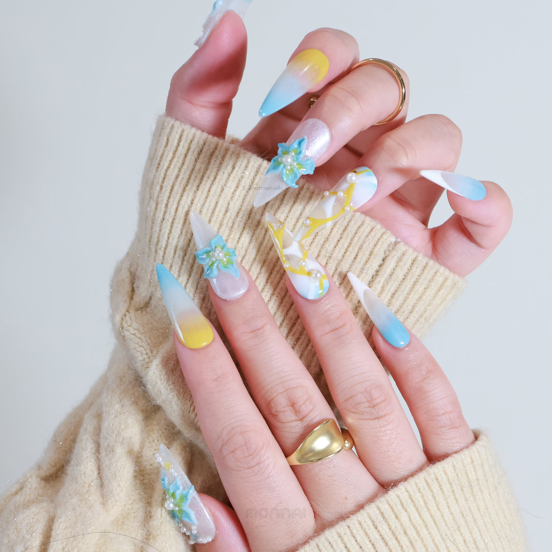 Ocean Breeze press-on nails shown on hands, with blue florals, pearls, and soft yellow-blue gradient in an ethereal, sculptural style