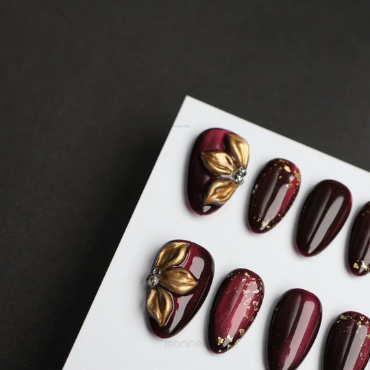 Opulent Noir press-on nails in deep burgundy with gold floral accents and scattered foil details.
