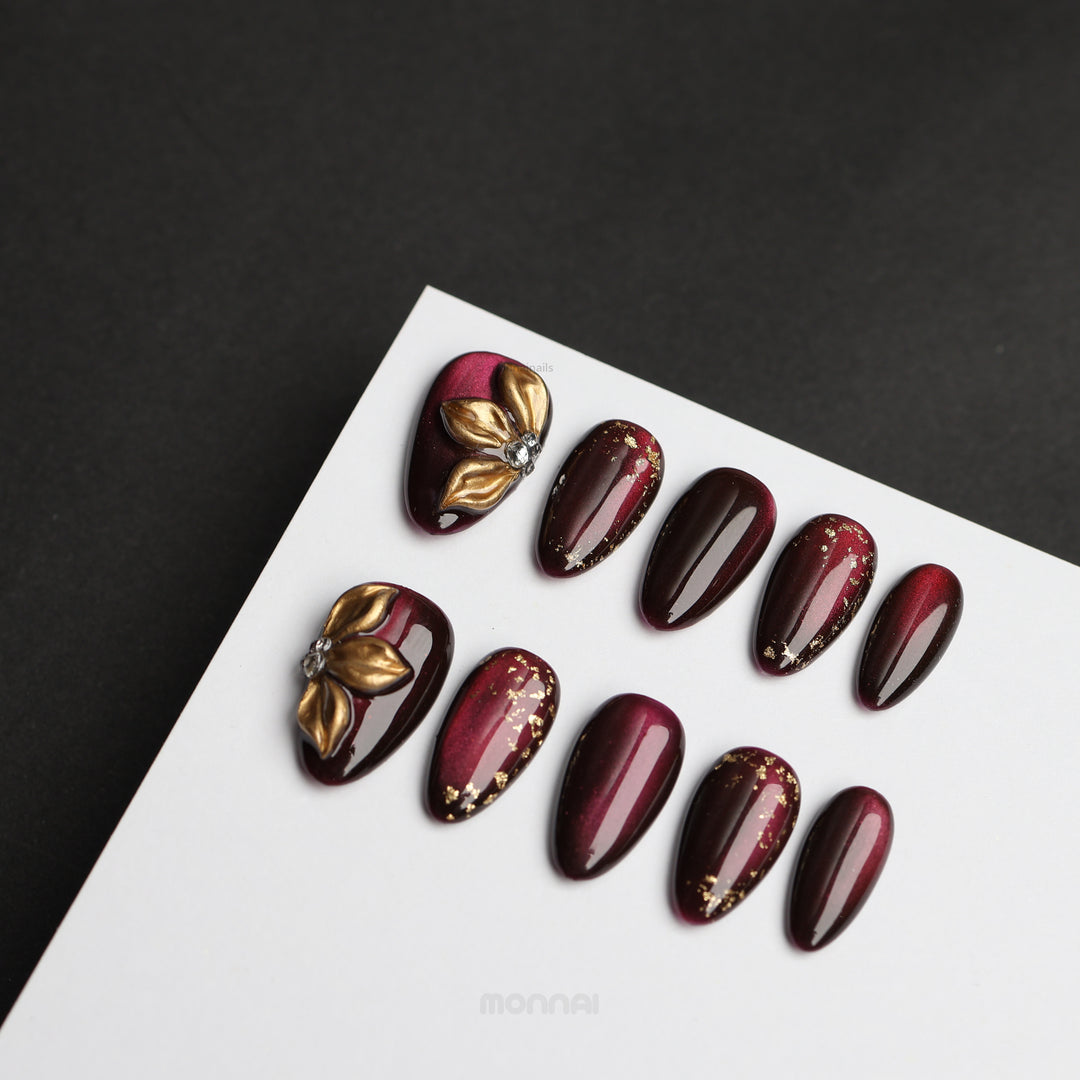 Opulent Noir press-on nails laid out with burgundy cat-eye shimmer, gold floral accents and foil details